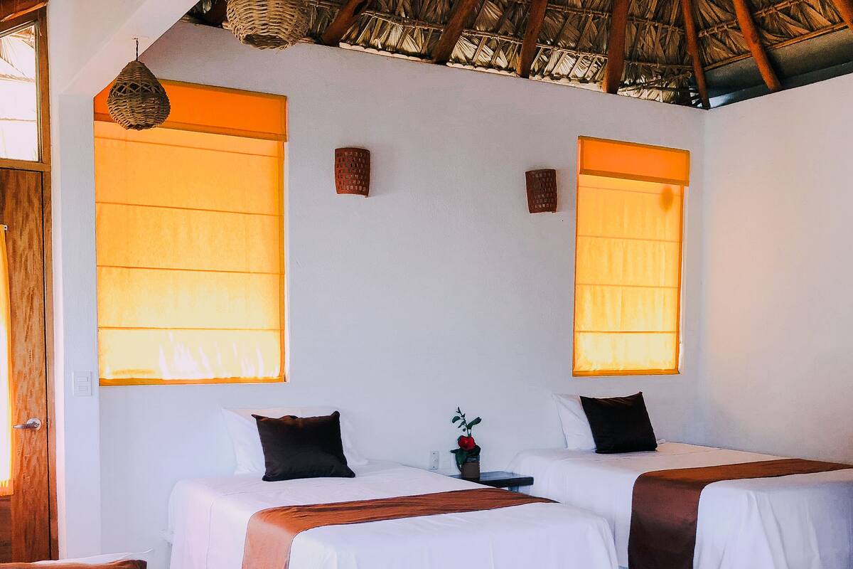 A room with two beds, each with white linens and brown pillows, a small table with a red flower, and a woven ceiling.