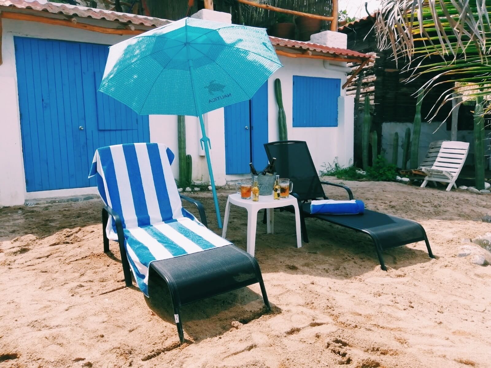 Een strandsetting met twee ligbedden, een gestreept en een effen, een klein tafeltje met drankjes en een blauwe parasol.