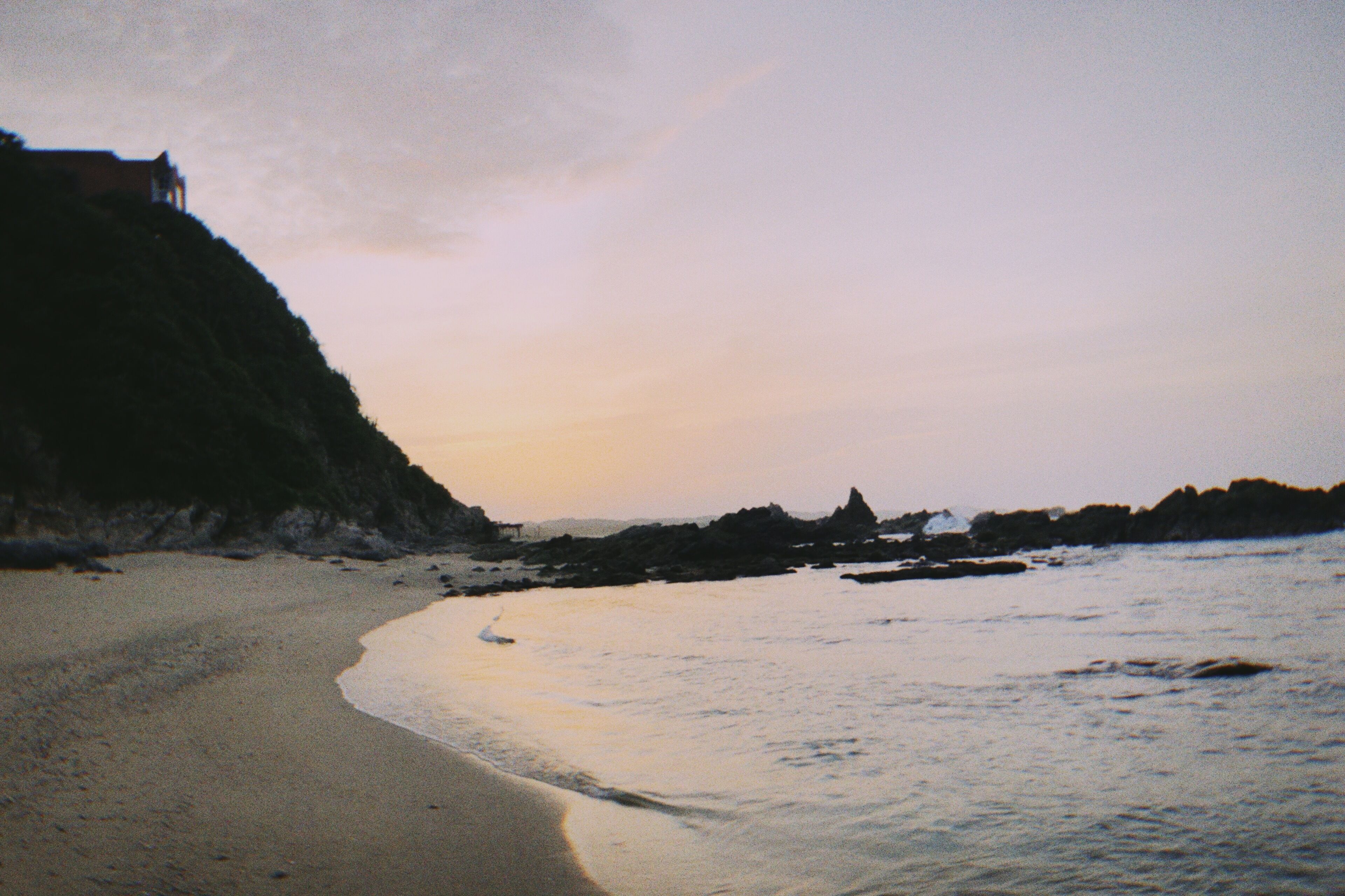 A beach with a building on a cliff, calm water, and rocky outcrops.