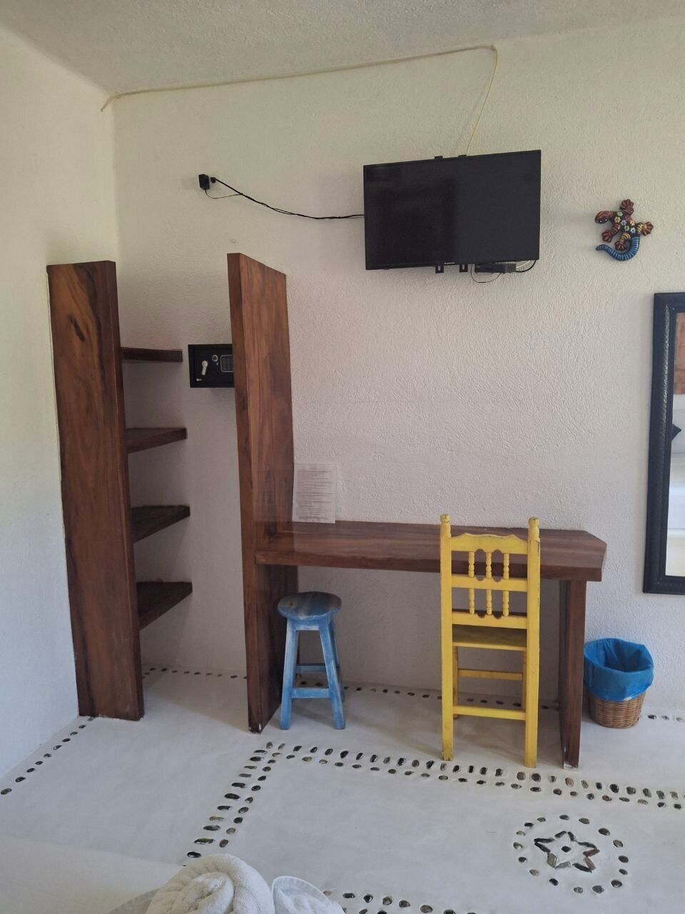 Una habitación con un escritorio de madera, un taburete azul, una silla amarilla, un televisor montado en la pared y un piso con diseño.