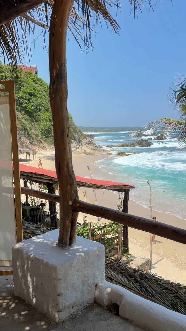 Vista de la playa con una estructura de madera, una palmera y un edificio al fondo.