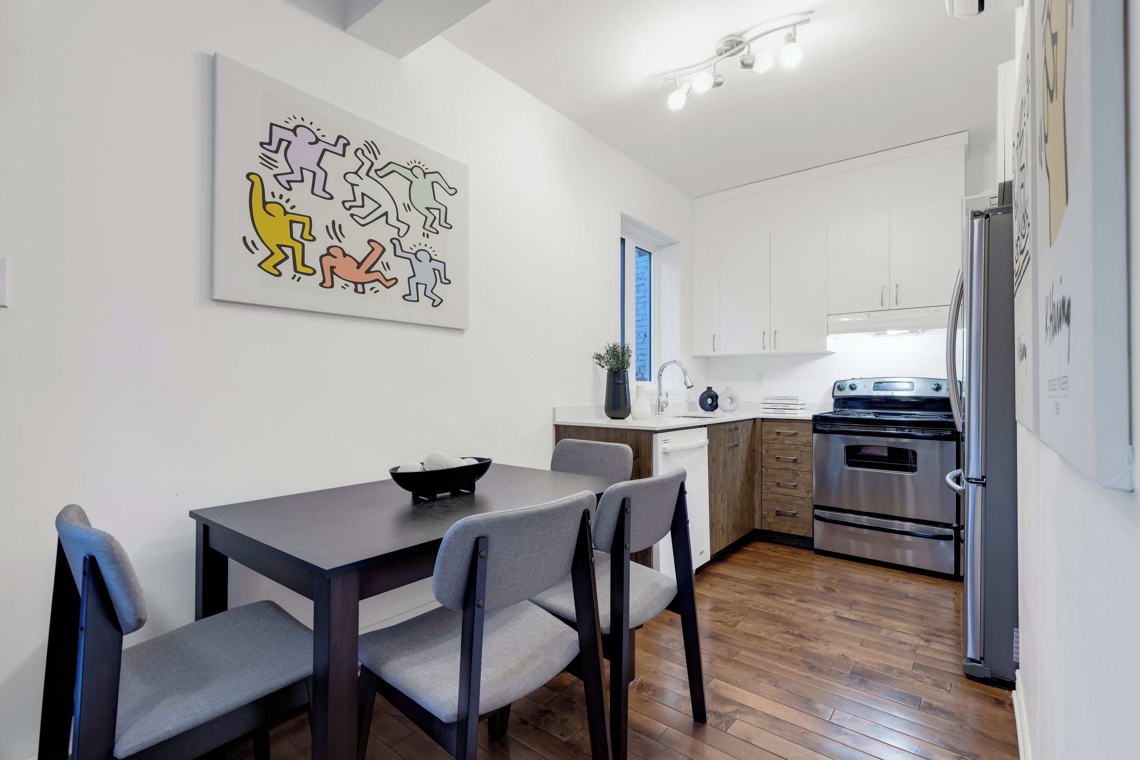 A modern kitchen with white cabinets, a stainless steel refrigerator, and a dining area with a dark table and grey chairs.