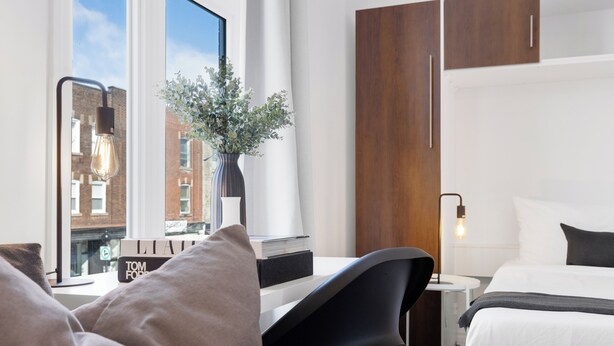 A modern hotel room with a large window, a desk with a vase of flowers, a bed with white linens, and a nightstand with a lamp. - LiV MTL - MTL (Montreal)