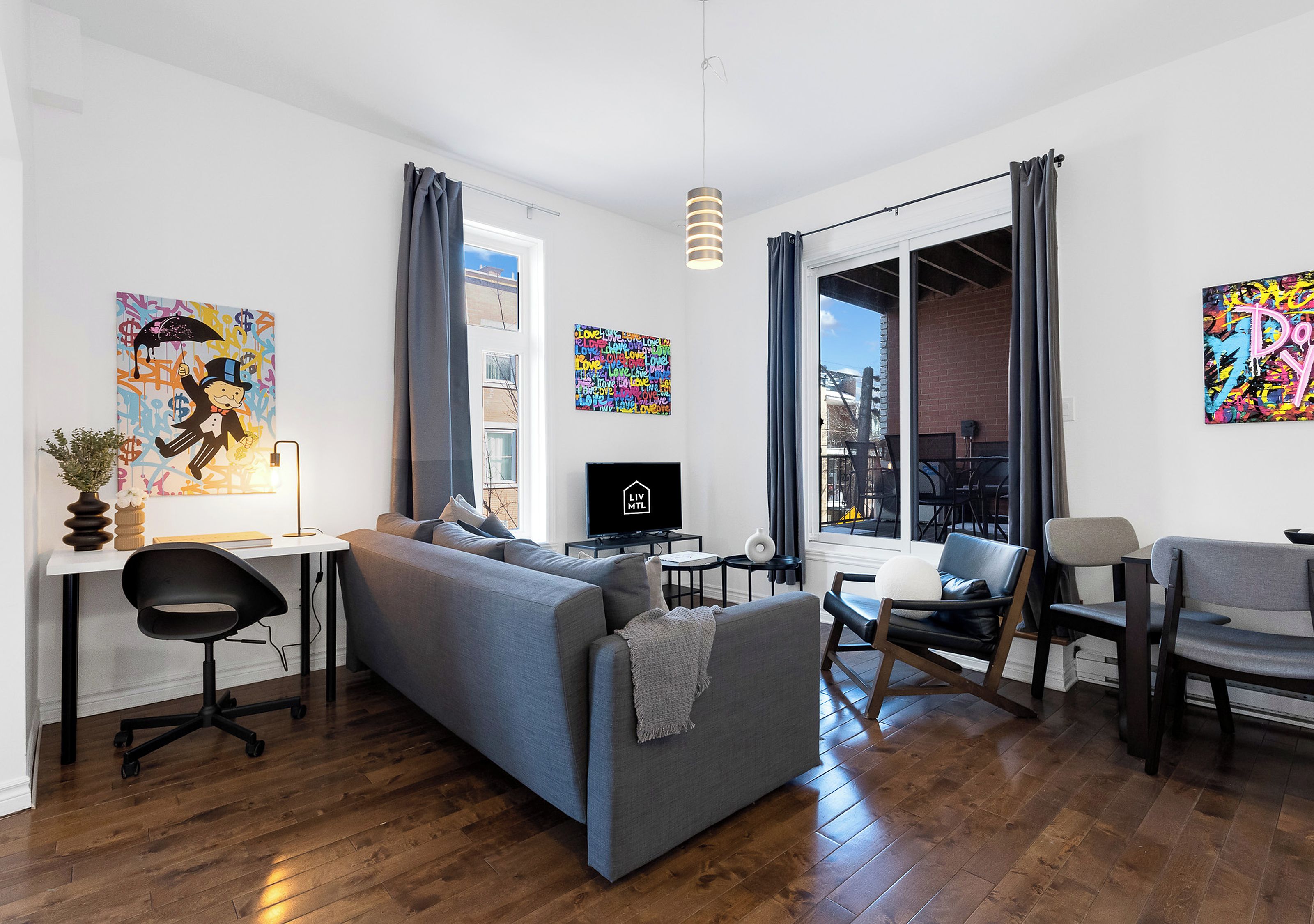 Una sala de estar moderna con un sofá gris, un escritorio con una computadora y un televisor. Hay dos sillones, una planta y obras de arte en las paredes.