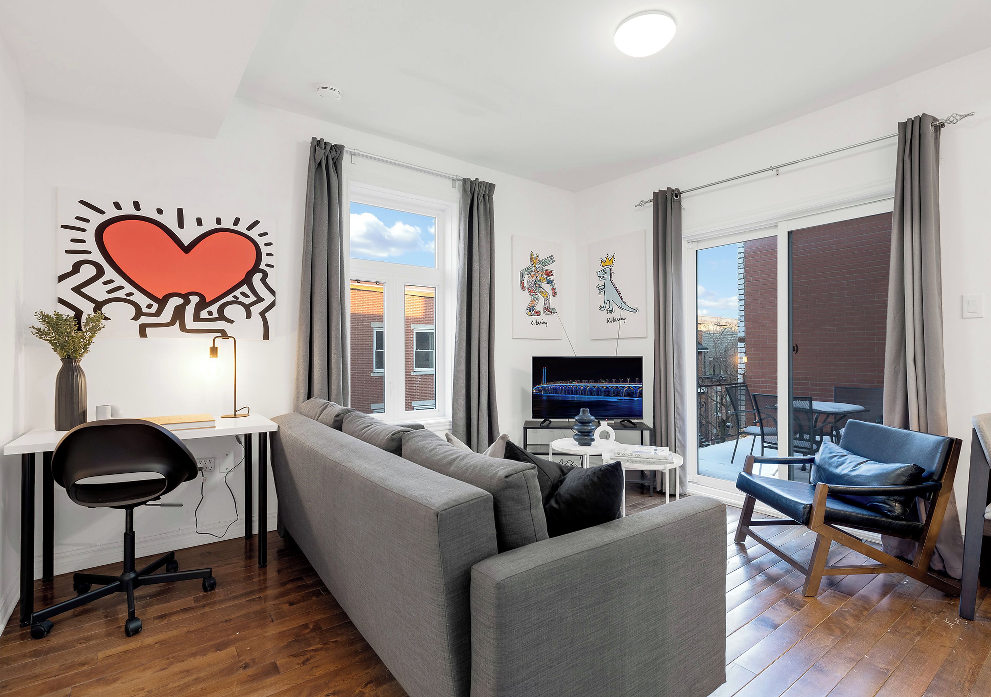 Un salon moderne avec un canapé gris, un fauteuil de bureau noir, un téléviseur et des œuvres d’art accrochées aux murs.