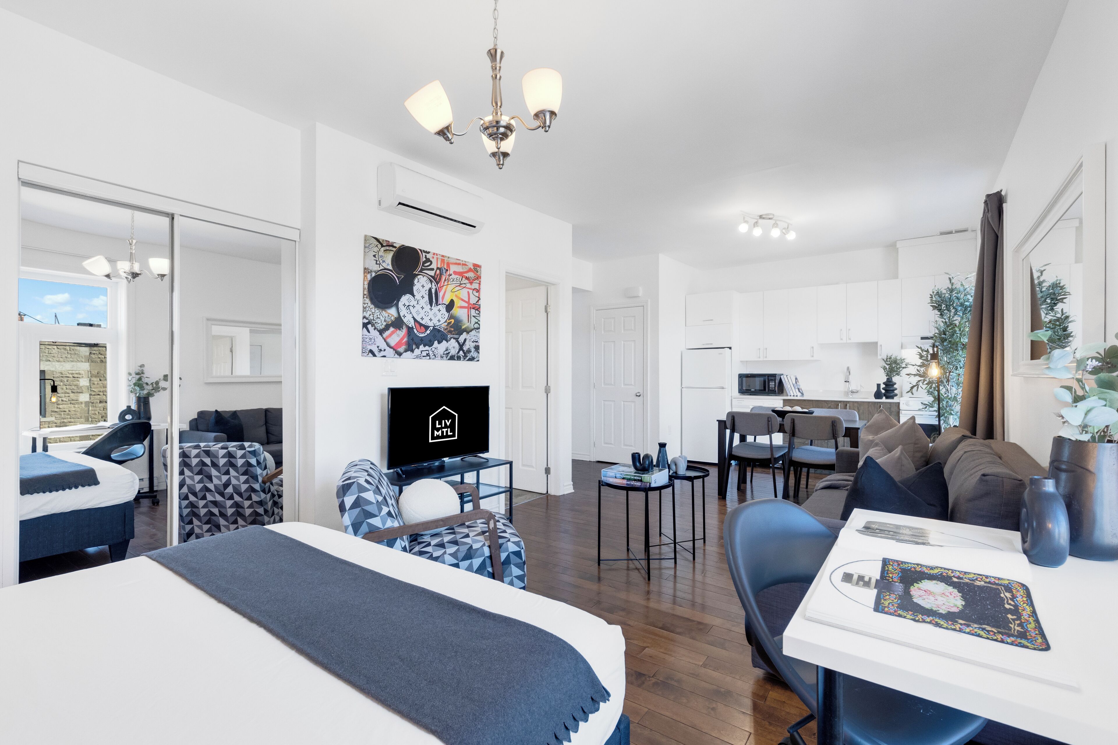 A modern hotel room with a bed, a desk, a dining area, and a kitchen.