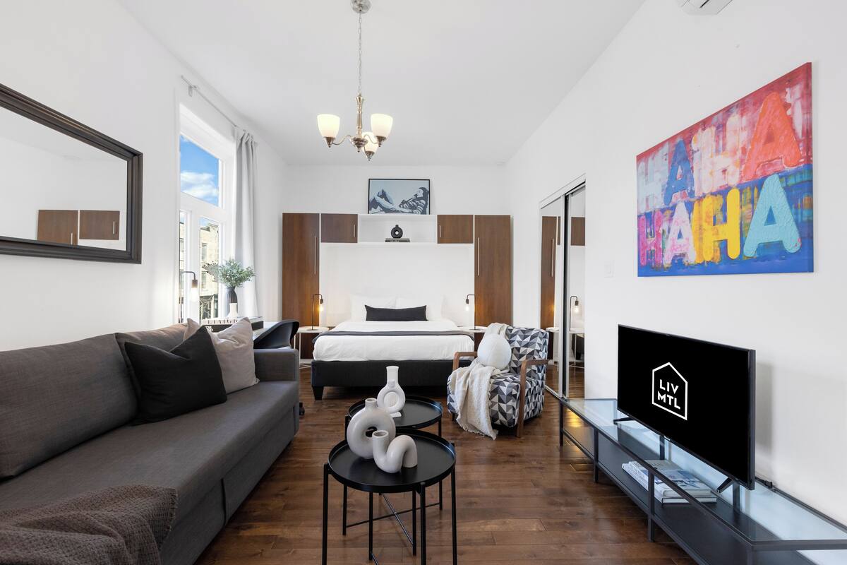 A modern hotel room with a bed, sofa, TV, artwork, and a chandelier.