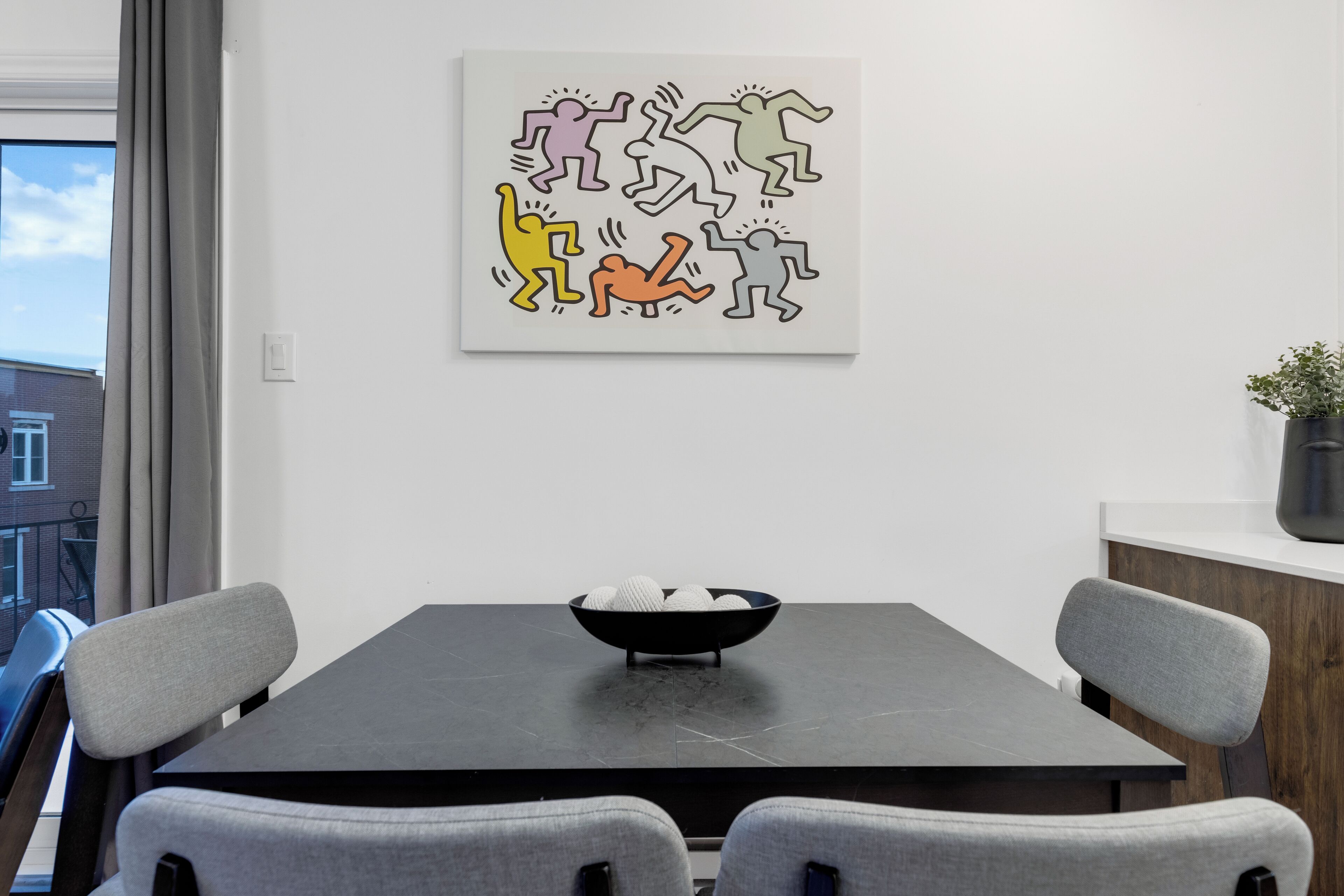A modern dining area with a dark table, grey chairs, and a bowl of white spherical objects. A colorful abstract artwork is on the wall.