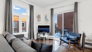 A modern living room with a grey sofa, a small white table, and a blue chair. There is a TV on a stand, a window with curtains, and artwork on the wall. - LiV MTL - MTL (Montreal)