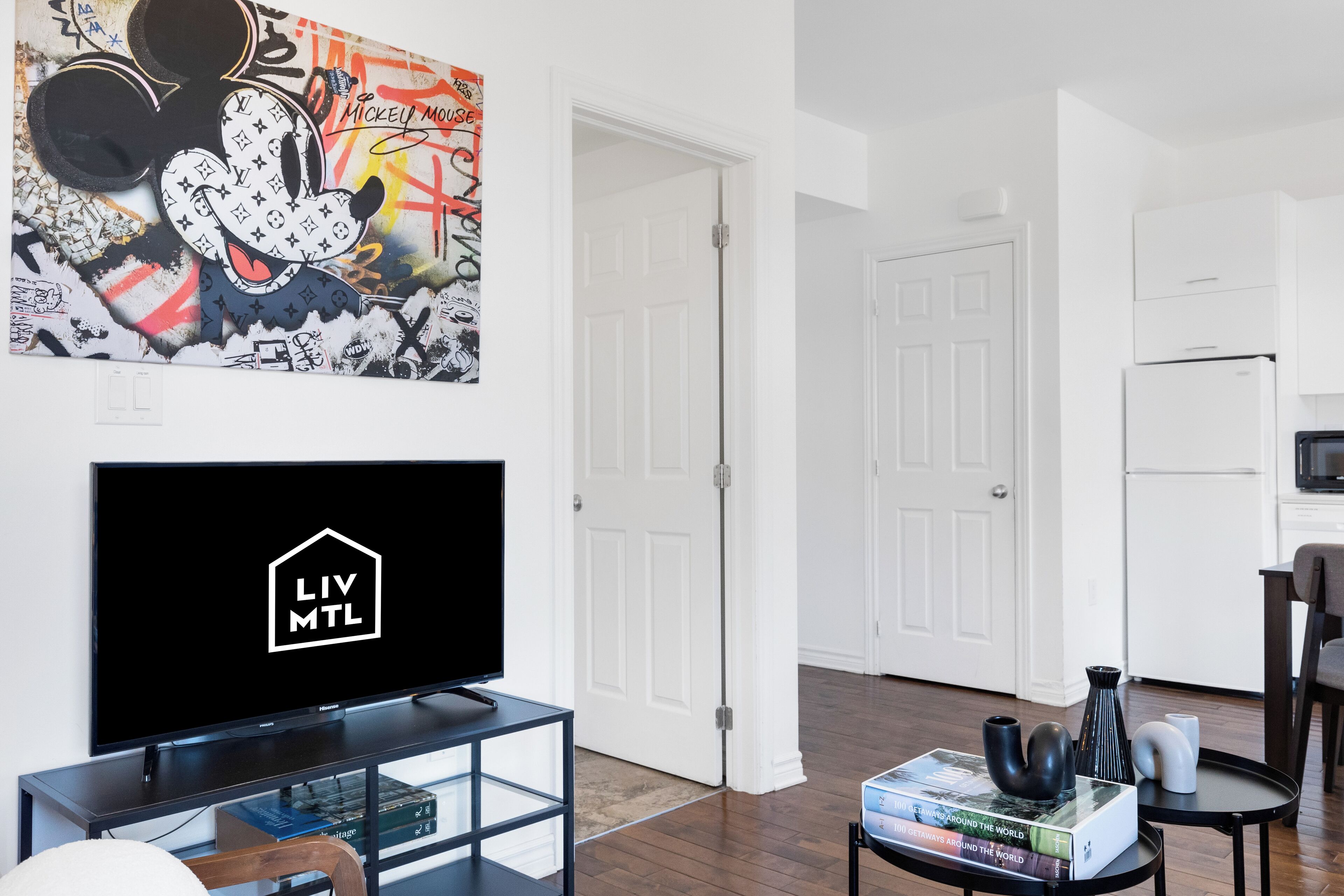 A modern living room with a flat-screen TV displaying a logo, a bookshelf, a coffee table with books and vases, and a large Mickey Mouse artwork on the wall.