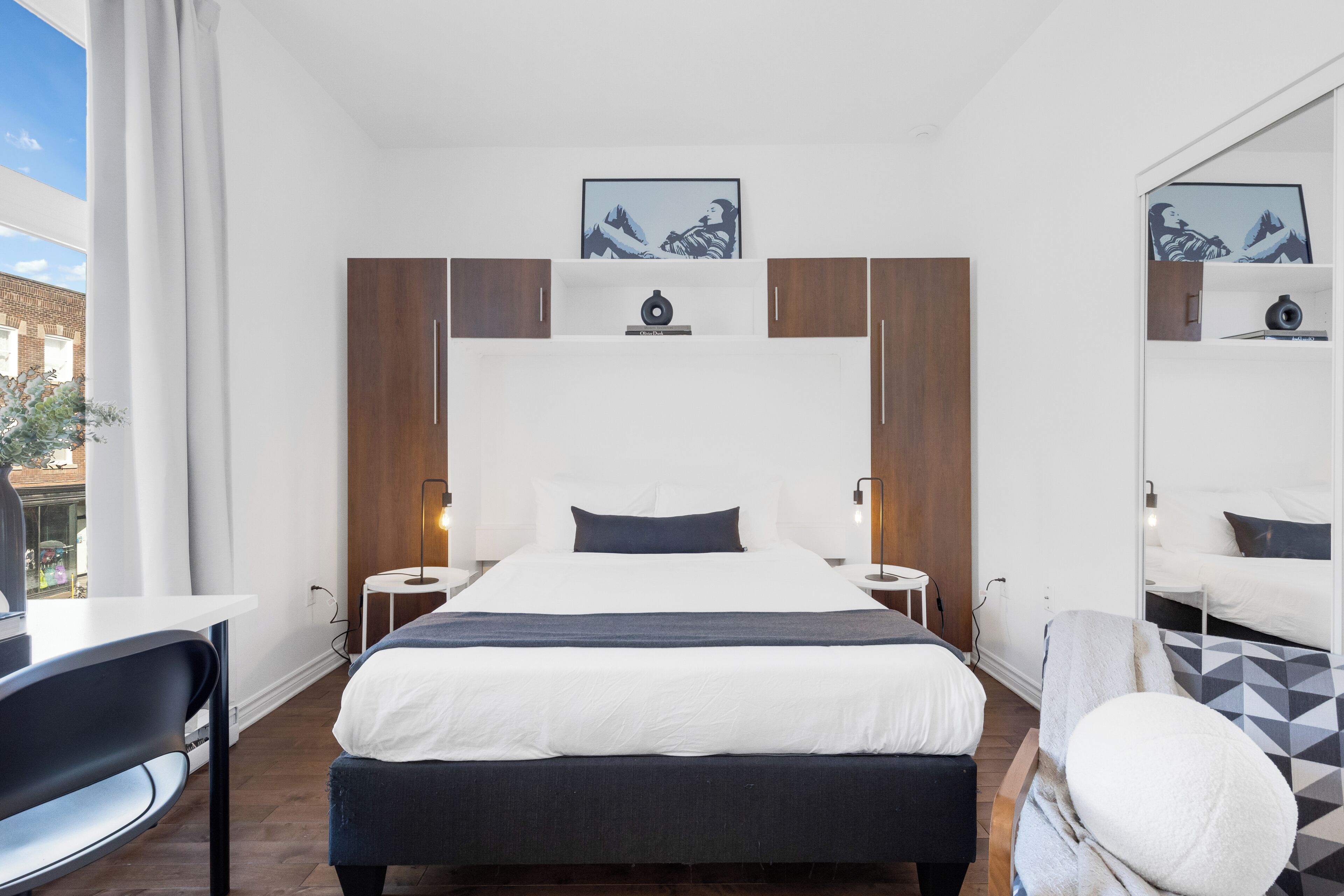 Una habitación de hotel moderna con una cama grande, un escritorio y una silla. Hay dos mesitas de noche con lámparas, un espejo grande y una obra de arte en la pared.
