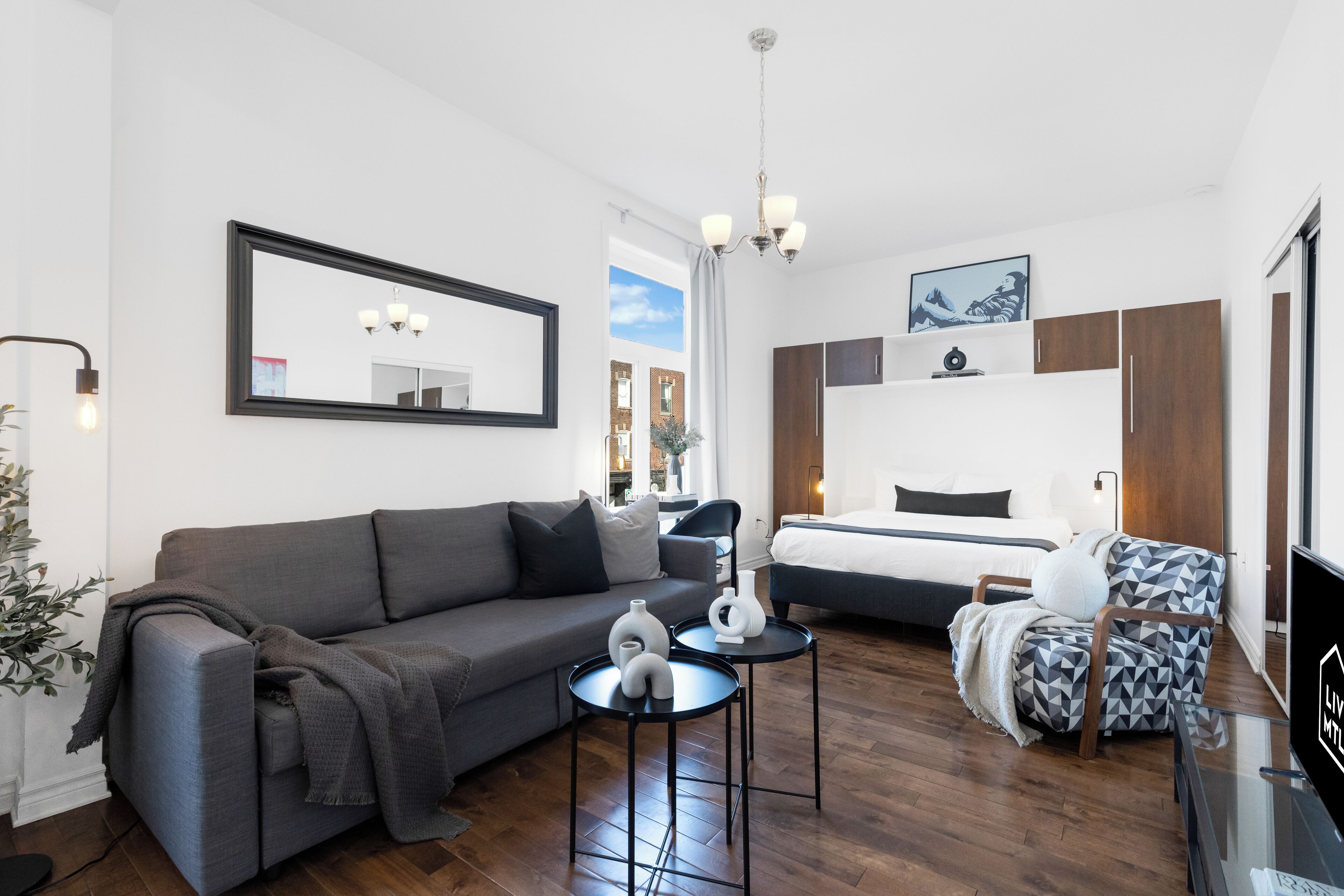 Moderni hotellihuone, jossa on harmaa sohva, sänky, kuviollinen nojatuoli, pieni pöytä koriste-esineineen ja televisio.
