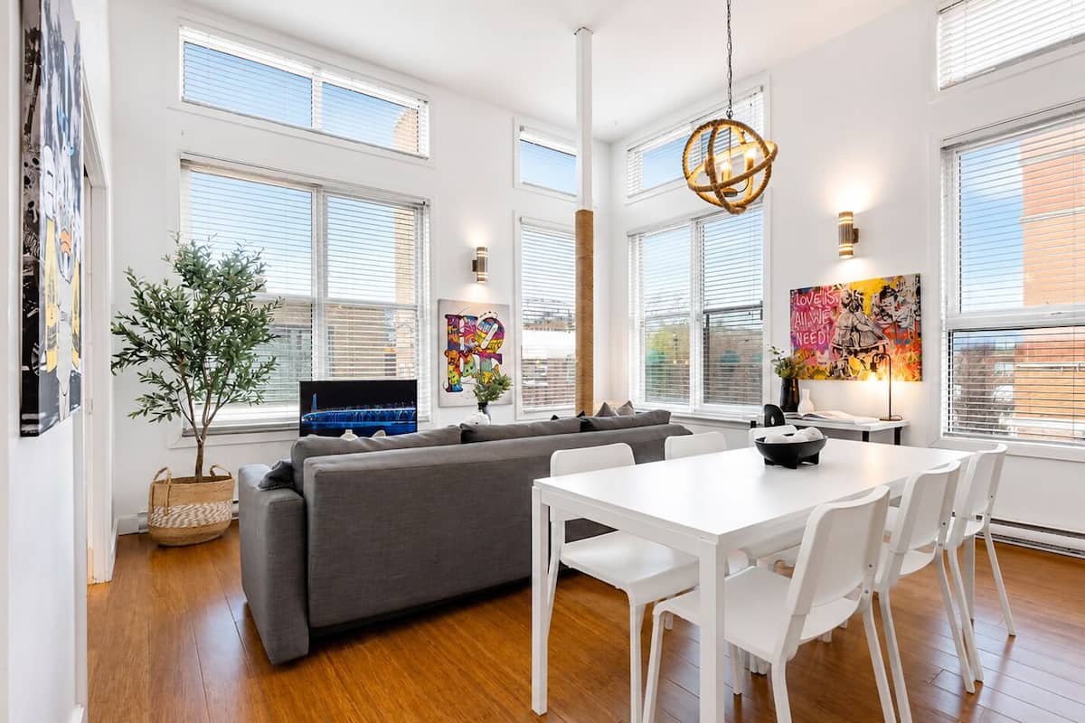 Una sala de estar moderna con un sofá gris, una mesa de comedor blanca y un ventanal con persianas.