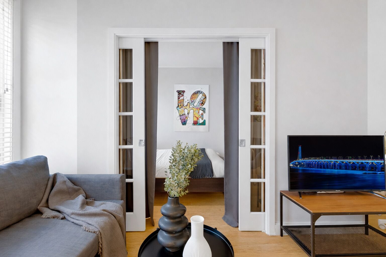 A modern living room with a sofa, a TV on a stand, a painting on the wall, and a door leading to a bedroom.