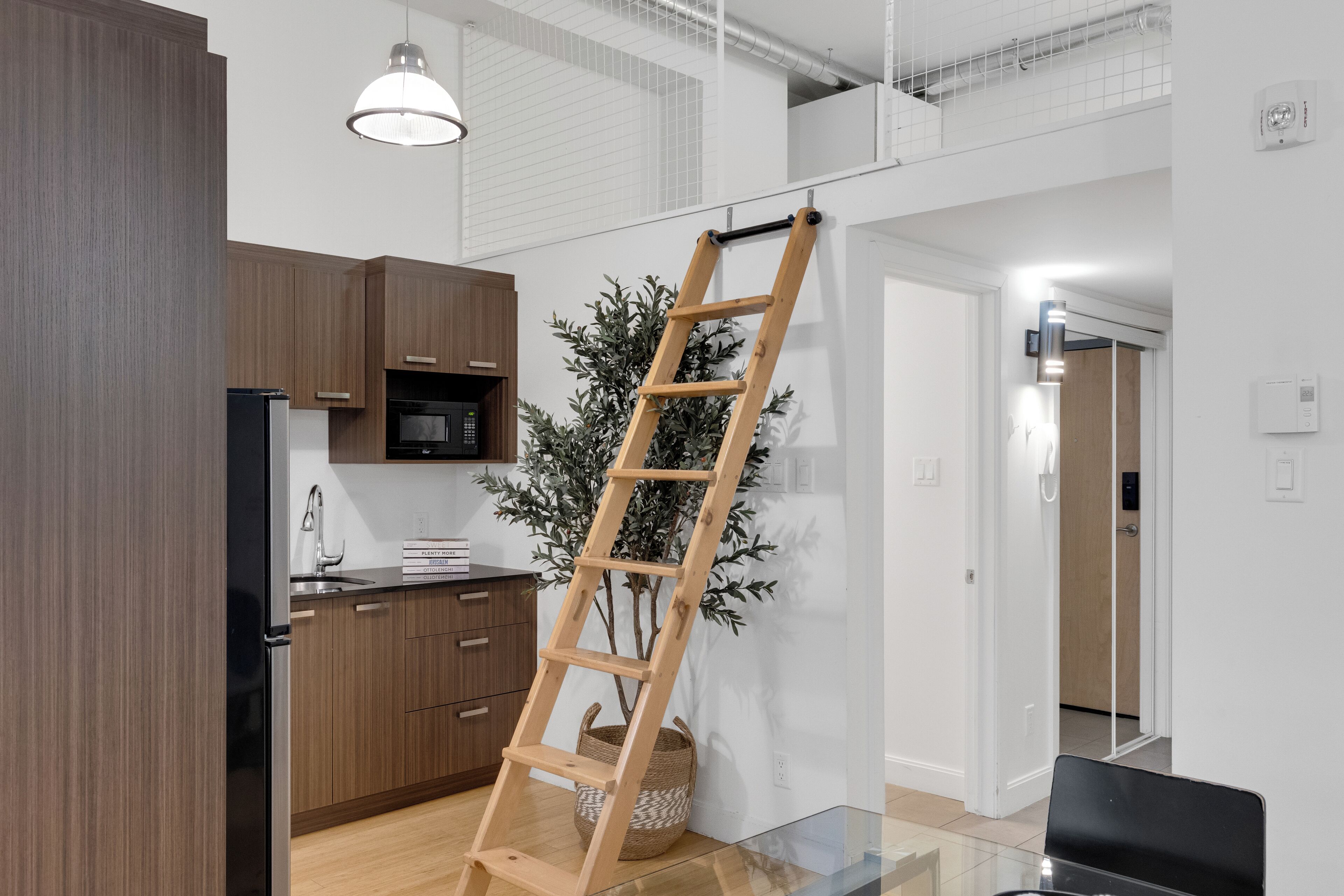 A modern kitchen with wooden cabinets, a stainless steel refrigerator, and a microwave. A wooden ladder is propped against the wall, and a potted plant is visible.