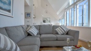 A modern living room with a grey sectional sofa, a small table with a glass of wine, and a large window with a view.