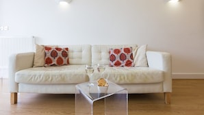 A white leather sofa with patterned cushions, a clear glass coffee table, and two wall-mounted lights. - Indigo (St Ives)