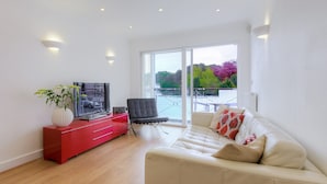 A modern living room with a red entertainment unit, a beige sofa, a black chair, and a large sliding glass door leading to a balcony with a view of trees and water. - Indigo (St Ives)