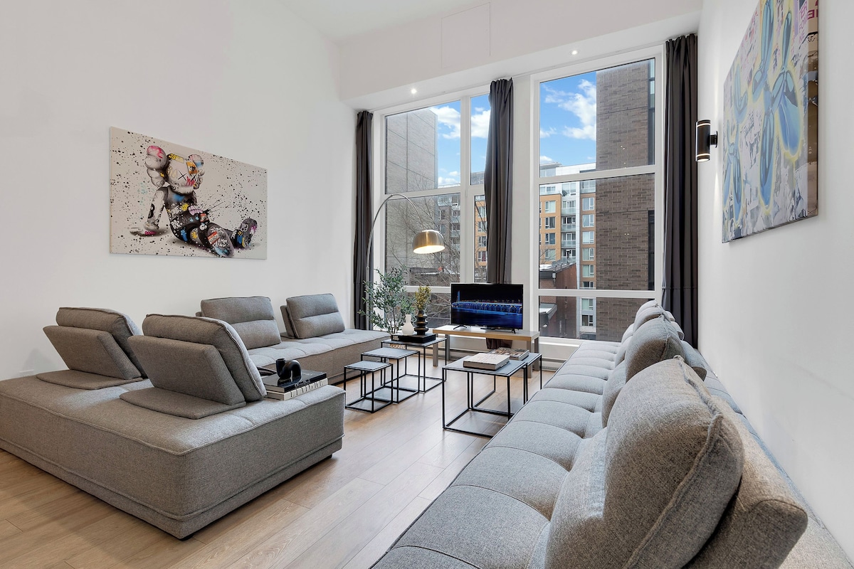 A modern living room with a grey sofa set, a flat-screen TV, and a large window with a city view.