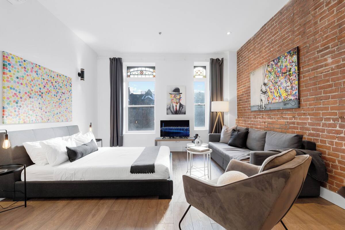 A modern hotel room with a bed, a sofa, a TV, and a brick wall.