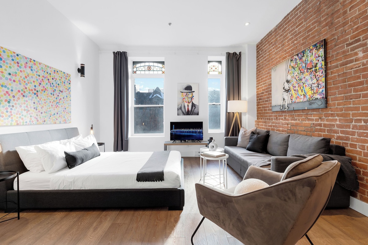 A modern hotel room with a bed, a sofa, a TV, and a brick wall with graffiti.
