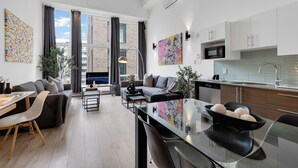 A modern apartment with a living room, dining area, and kitchen. It features a large window, a sofa, a dining table, and a kitchen with a microwave and sink. - LiV MTL - Downtown (Montreal)