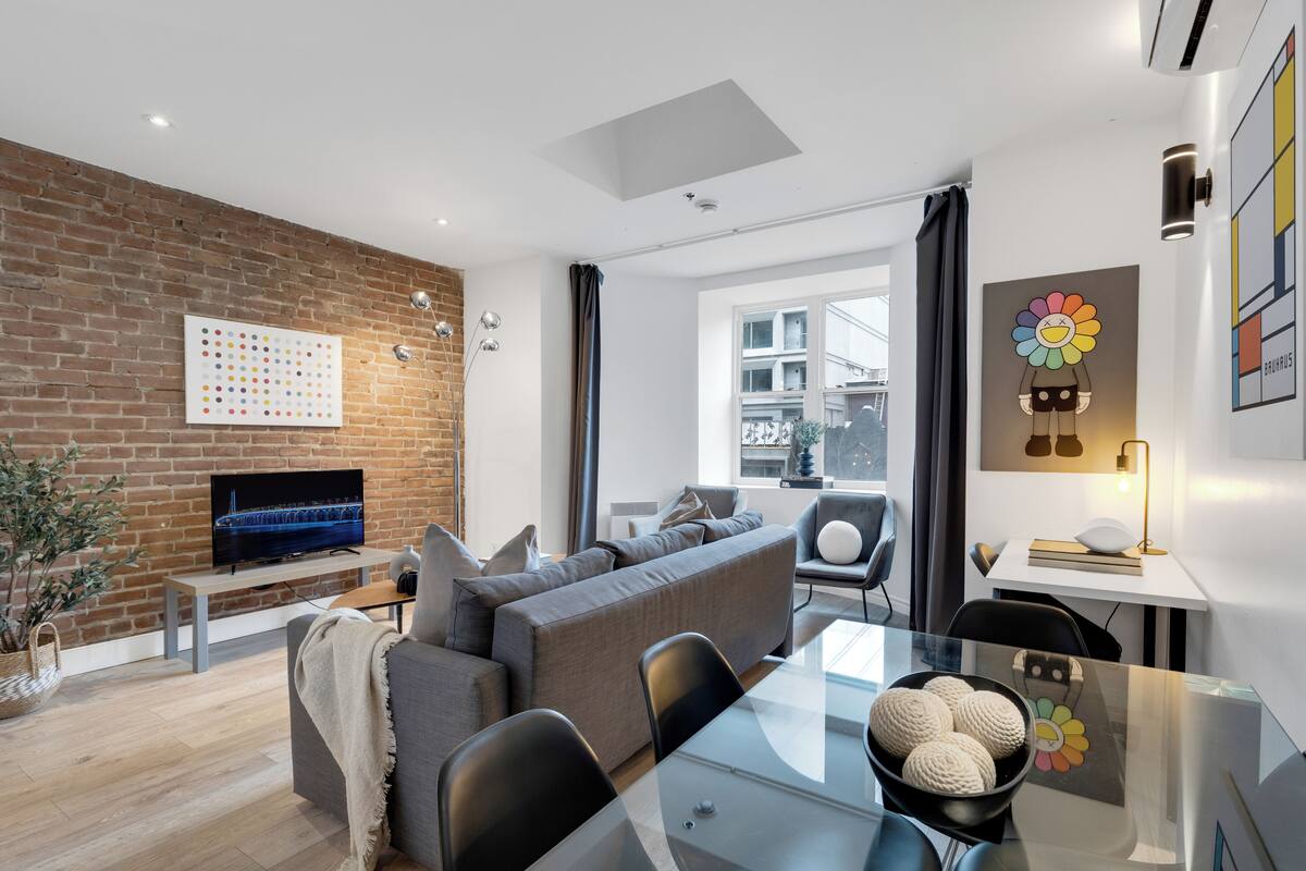 A modern living room with a brick wall, a flat-screen TV, a grey sofa, a glass dining table, and a window with curtains.