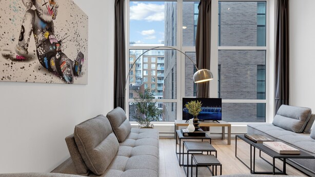 A modern living room with a grey sofa, a coffee table, a TV, and a large window with curtains. - LiV MTL - Downtown (Montreal)
