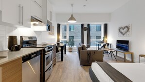 A modern apartment with a kitchen, dining area, and living room. It includes a dishwasher, oven, and a large window with a view of the city. - LiV MTL - Downtown (Montreal)
