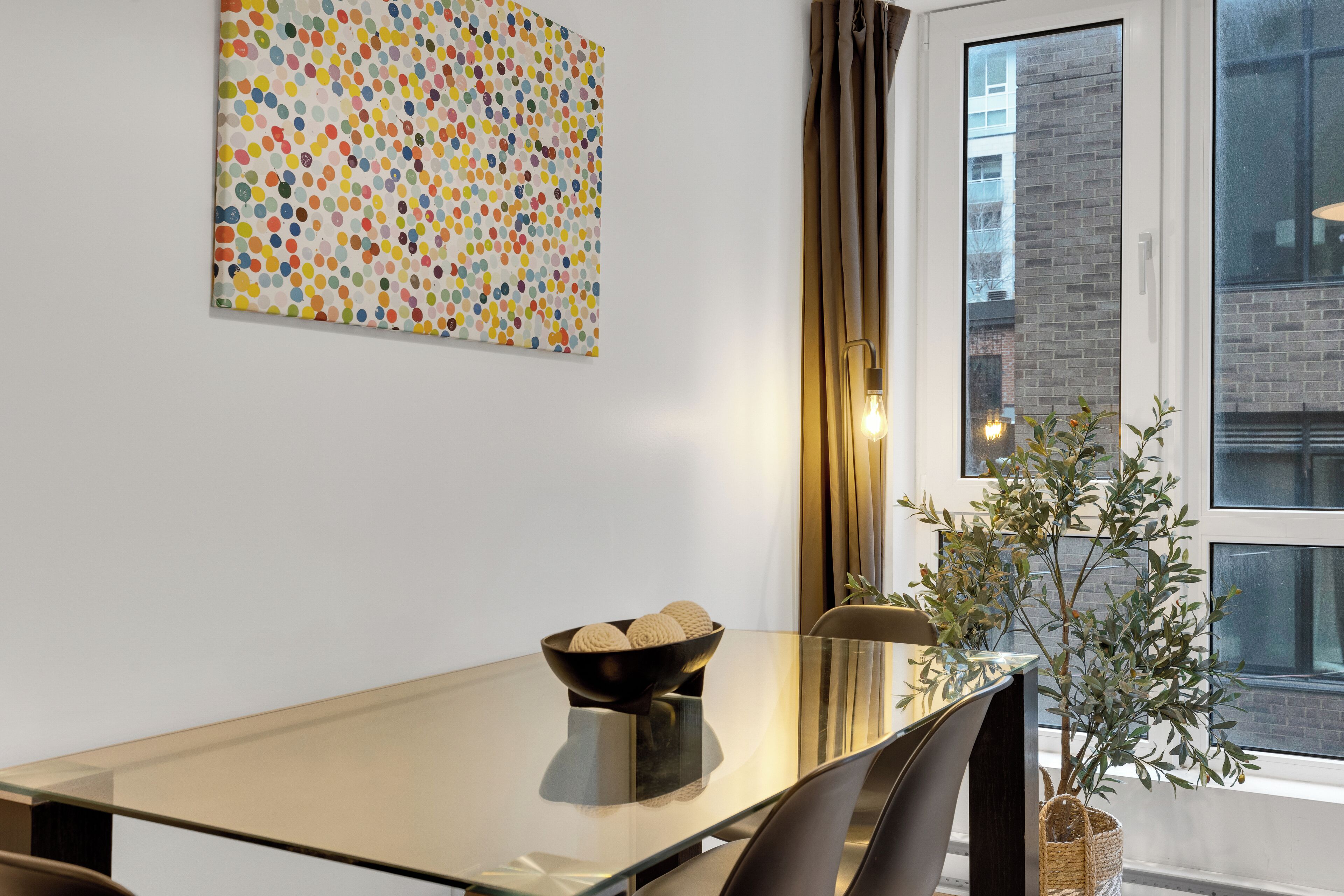 A modern dining area with a glass table, a bowl of decorative items, a colorful dot-patterned artwork on the wall, and a view of a building through a window.