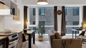 A modern kitchen with a built-in oven, a dining area with a table and chairs, a sofa, and large windows with curtains. - LiV MTL - Downtown (Montreal)