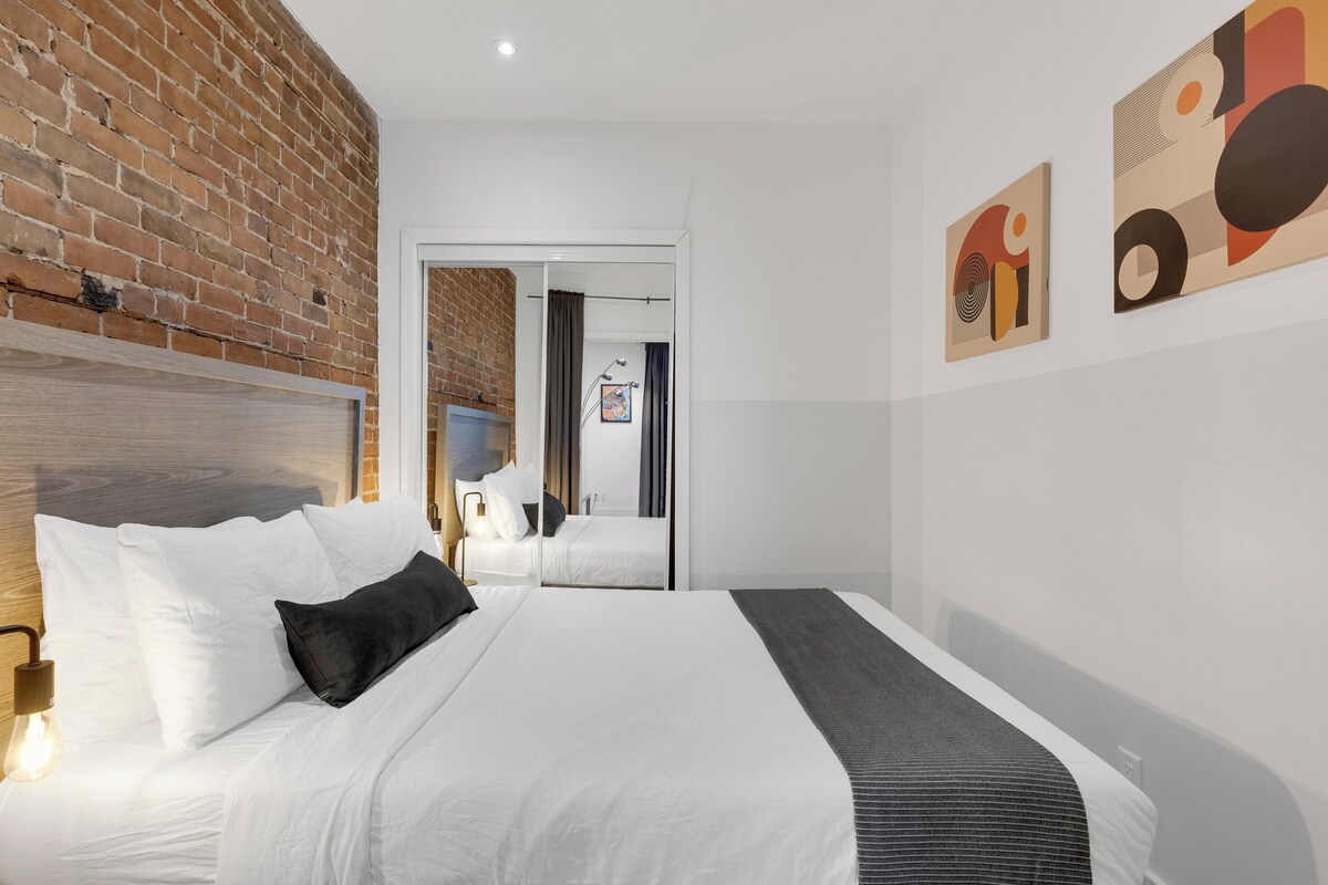 A modern hotel room with a brick wall, a large bed with white bedding, a headboard, and a bedside table with a lamp. There are abstract paintings on the wall and a door leading to another room.