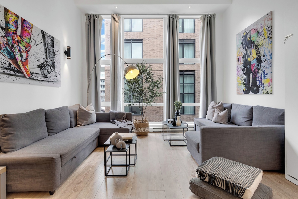 A modern living room with grey sofas, a coffee table, and a floor lamp. There are large windows with curtains, a potted plant, and artwork on the walls.
