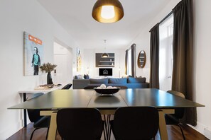 Un espace de repas moderne avec une longue table, des chaises noires, un récipient contenant des objets décoratifs, un luminaire suspendu et un canapé en arrière-plan.