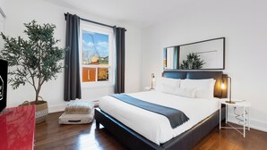 Une chambre à coucher avec un lit, une fenêtre avec des rideaux, une table de chevet avec une lampe, une plante en pot et une chaise rouge.