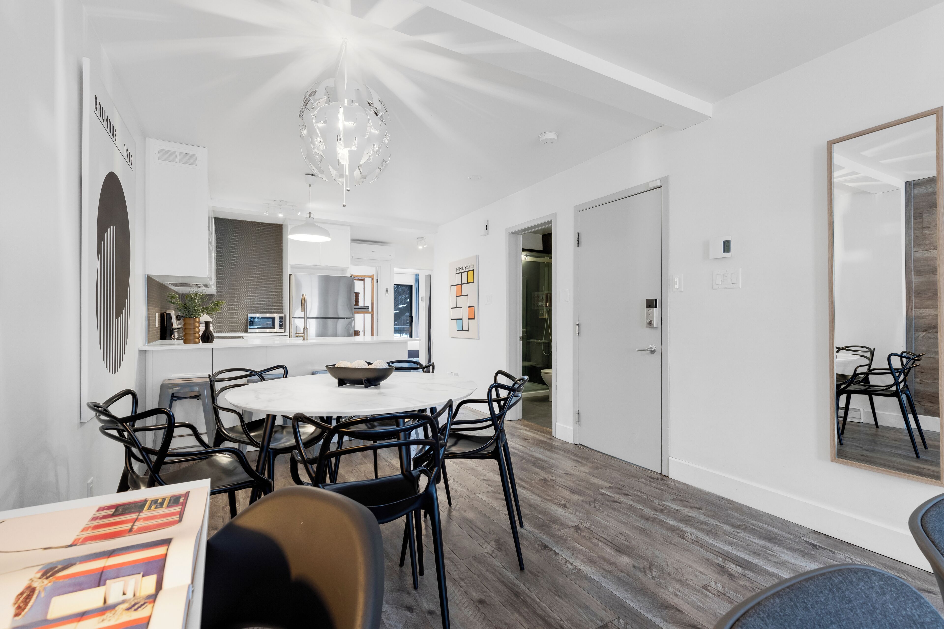 A modern dining area with a round table and chairs, a kitchen in the background, and a large mirror on the wall.