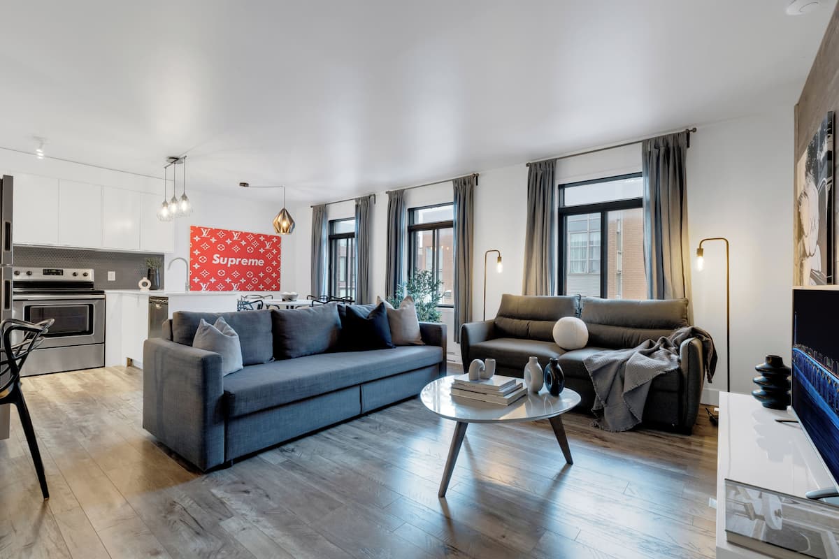 Een moderne woonkamer met een grijze bank, een salontafel en een televisie. Er is een keuken met een fornuis en kastjes, en een rood bord met de tekst 'Supreme' aan de muur.