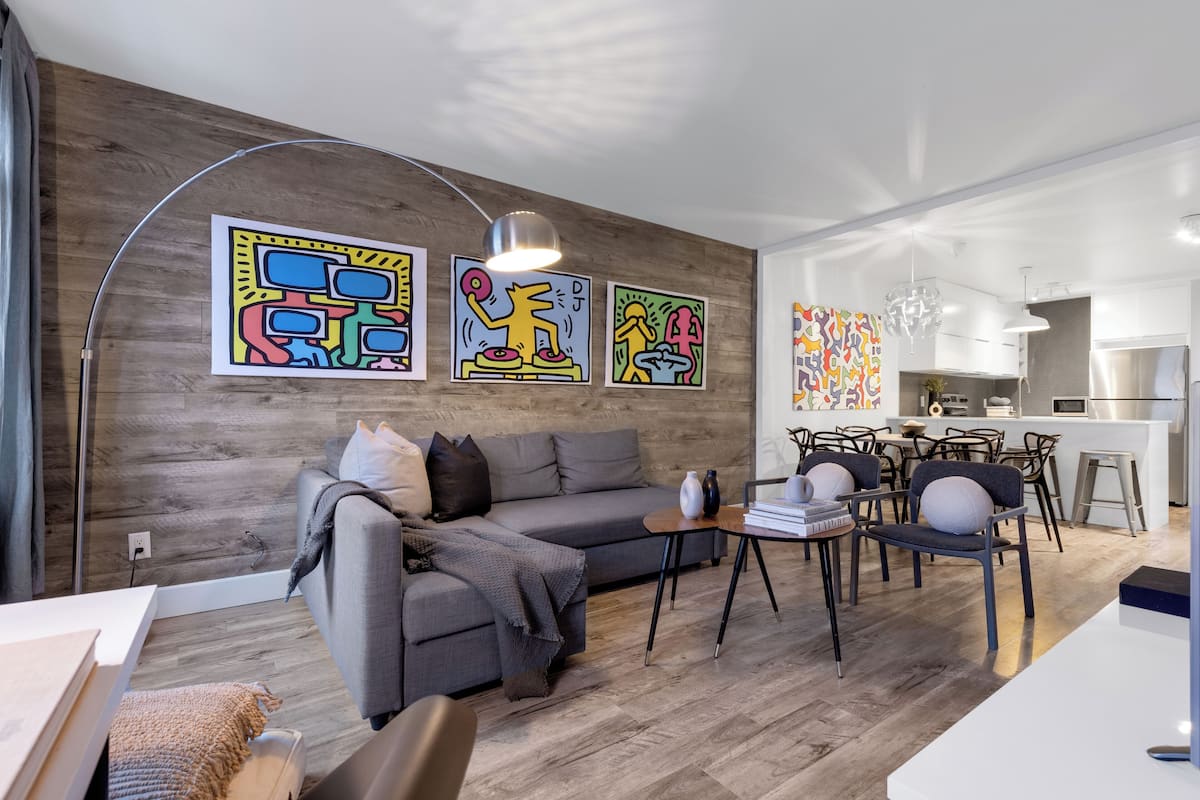 Een moderne woonkamer met een grijze bank, een salontafel, en een eethoek met stoelen en een tafel. Op de muur hangen kleurrijke abstracte schilderijen en er is een keuken te zien op de achtergrond.