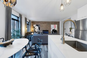 A modern kitchen with a stainless steel refrigerator, a white countertop, and a built-in oven. There is a dining area with a white table and chairs, and a sofa in the background. - LiV MTL - Urban (Montreal)