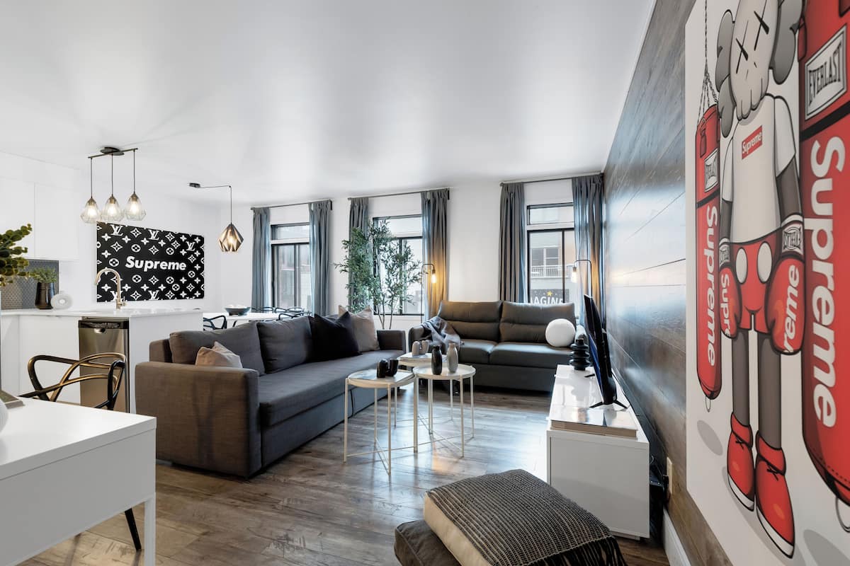 Een moderne woonkamer met een grijze bank, een witte salontafel en een tv op een meubel. Op de muur een groot muurdecor met het woord 'Supreme' en een afbeelding van een personage.