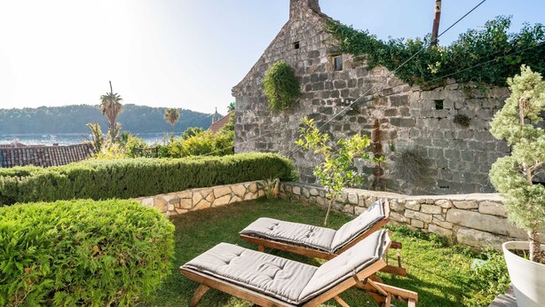 A stone building with a sloped roof, a small window, and a chimney. There are two wooden lounge chairs with cushions. The building is surrounded by well-maintained hedges and a small tree. - Villa Vista Old Town (Konavle)