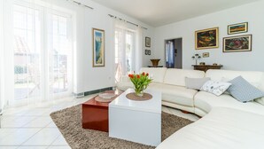 A modern living room with a white sofa, a coffee table with a vase of flowers, and a large window with sheer curtains. - Villa Vista Old Town (Konavle)