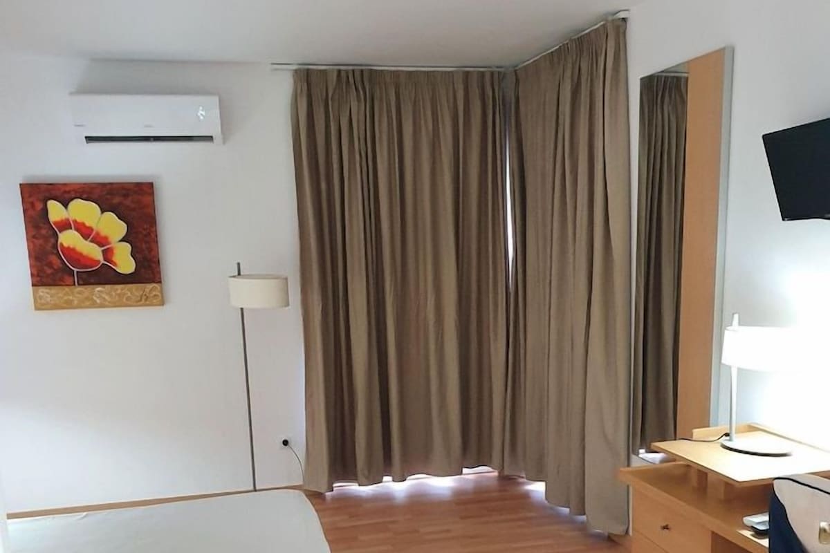 Quarto de hotel com cama, escrivaninha, cadeira, televisão e janela com cortinas.