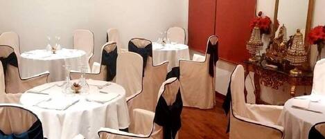 Un comedor con mesas redondas, sillas adornadas con cintas negras y una pared roja con un espejo y elementos decorativos.