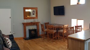 A living room with a sofa, a fireplace, a dining table with chairs, a TV, and a kitchen area with a microwave. - Apartment Sleeps 7 With Mezzanine (Birr)