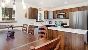 A modern kitchen with wooden cabinets, stainless steel appliances, a dining area with a wooden table and chairs, and a hanging light fixture. - Hawkeye Ridge -- Ev 6189 3 Bedroom Condo by RedAwning (Estes Park)