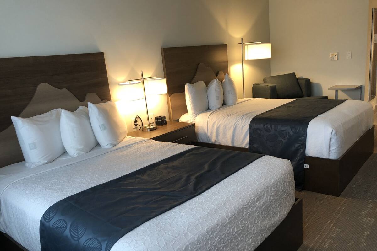 A hotel room with two beds, each with a headboard, bedside tables, lamps, and a small shelf.