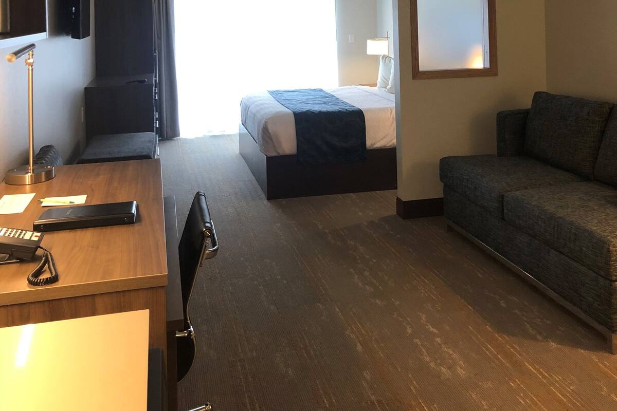 A hotel room with a bed, a desk with a telephone, a chair, a sofa, a TV mounted on the wall, and a window with curtains.