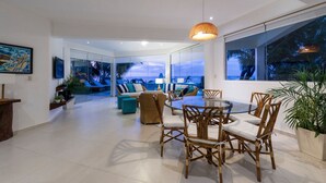 Amplia sala de estar con pared de vidrio que da a un balcón, una mesa redonda de vidrio con sillas y una lámpara colgante.