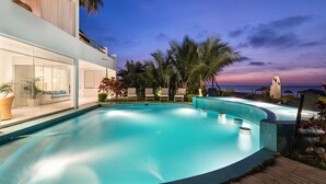Un área de piscina moderna con vista al océano al atardecer, rodeada de palmeras y sillones de descanso.